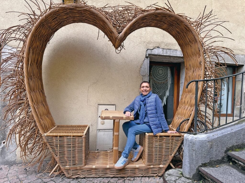 Sculpture en rotin en forme de cœur géant avec femme souriante assise à l'extérieur, décor naturel et ambiance chaleureuse.