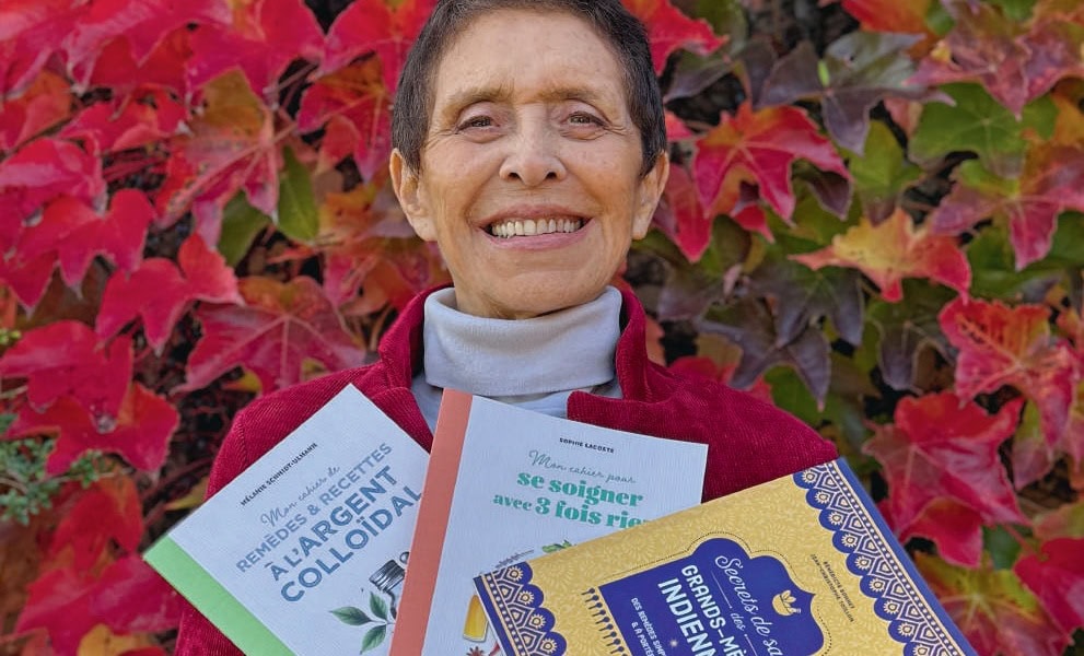 Souhaite une femme âgée avec des livres sur la santé et le bien-être, devant un mur de feuillages d'automne rouges et verts, pour promouvoir le contenu de Rebelle Santé.