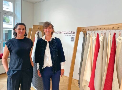 Deux femmes discutent dans un espace d'exposition de textiles naturels, avec des échantillons de tissus colorés suspendus, illustrant l'engagement pour des matériaux authentiques et durables.