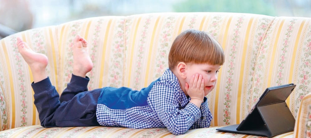Bébé allongé sur un canapé, regarde une tablette numérique avec curiosité et amusement, dans un intérieur lumineux.