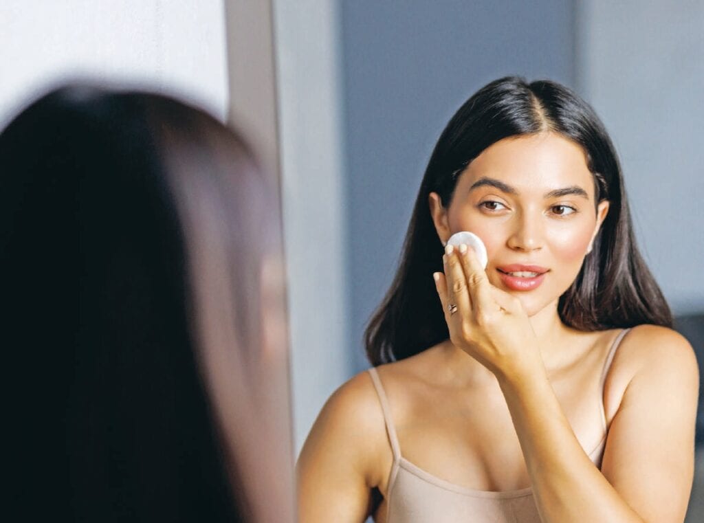 Jeune femme appliquant une crème sur sa joue dans un miroir, illustrant soin de la peau et beauté.
