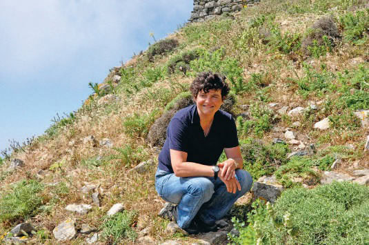 Femme souriante en pleine nature, assise sur un terrain en pente avec des plantes et des rochers, illustrant un moment de détente et de réflexion dans un environnement naturel.